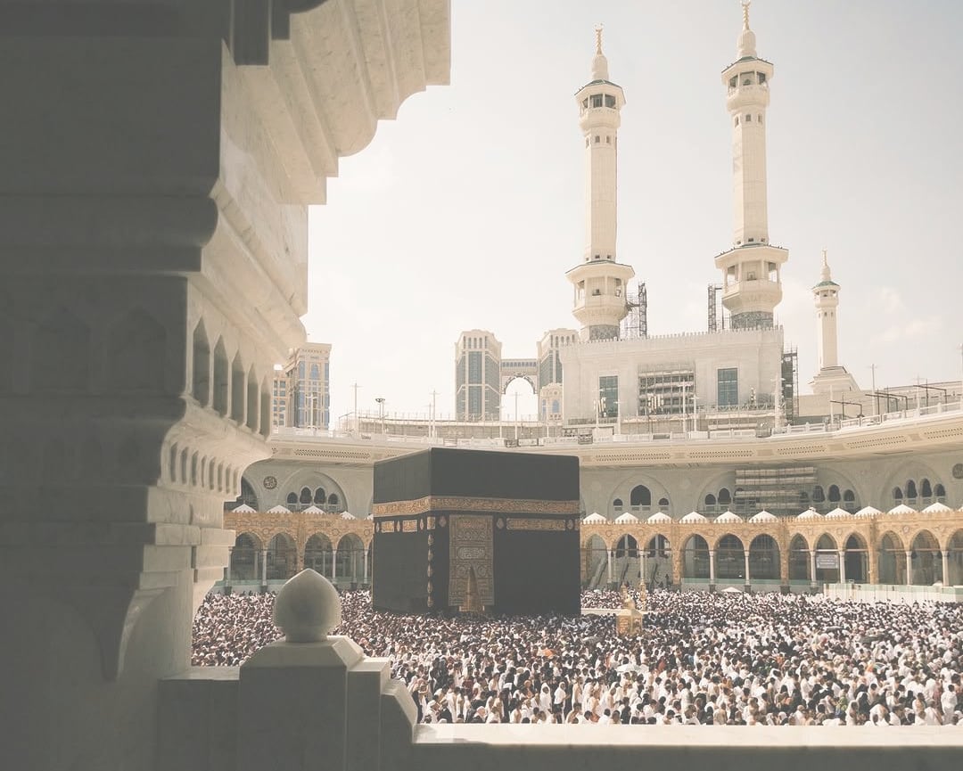 The Kaaba in the Grand Mosque at Makkah, seen past the columns of a shaded marble arcade, with pilgrims filling the mataf and the minarets of the Haram rising behind